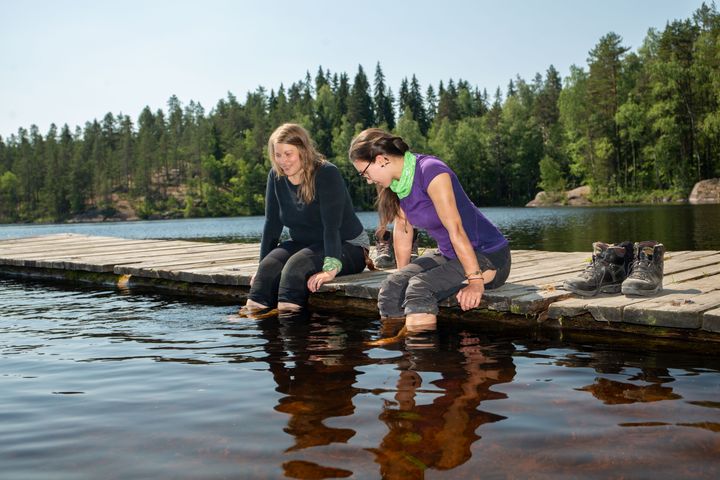 Kaksi naista istuu laiturilla ja uittaa jalkojaan vedessä. Taustalla on metsämaisema ja naisten vieressä laiturilla vaelluskengät.