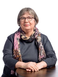 Päivi Rissanen hymyilee. Hänellä on lyhyet harmaanruskeat hiukset, silmälasit, harmaa paita ja kevyt kuviollinen kaulahuivi.