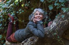 Irina Björklun hymyilee maatessaan puunoksalla ja nojaa leukaa kämmeneensä. Vihreät lehdet laskeutuvat köynnöksinä takana olevan seinämän päältä.