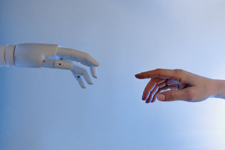 valkoinen robotin käsi ojentuuu vasemmasta laidasta kohti oikealta tulevaa ihmisen kättä, vaalea tausta