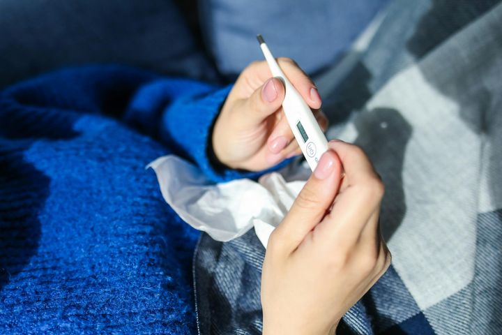 sinipaitainen henkilö lepää viltin alla, kuvassa näkyy vain keskivartalo, käsissä kuumemittari