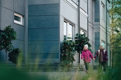Kaksi pientä lasta kävelee käsi kädessä hymyillen modernin rakennuksen ohi, jossa on tummat ja vaaleat paneelit seinässä. Lapset ovat pukeutuneet ulkovaatteisiin. Taustalla näkyy rakennuksen ikkunoita ja vihreitä kasveja.