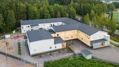 Ilmakuva suuresta L-muotoisesta koulurakennuksesta, jonka ympärillä on leikkipiha ja metsää.