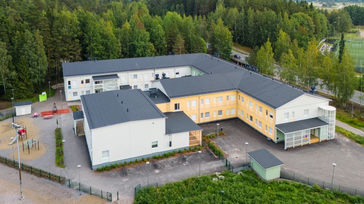 Ilmakuva suuresta L-muotoisesta koulurakennuksesta, jonka ympärillä on leikkipiha ja metsää.