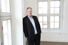 Mies seisoo sisätilassa ikkunan ääressä ja hymyilee. Hänellä on yllään tumma puku ja vaalea kauluspaita.