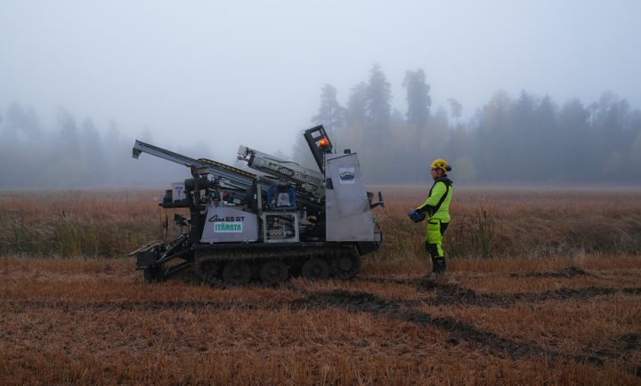 Arbetare i gul-grön varselkläder och hjälm står bredvid en bandgående borrmaskin på ett dimmigt, höstbrunt fält med skog i bakgrunden.