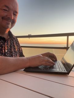 Henkilö hymyilee kameralle. Läppäri pöydällä ulkona, taustalla auringonnousu.