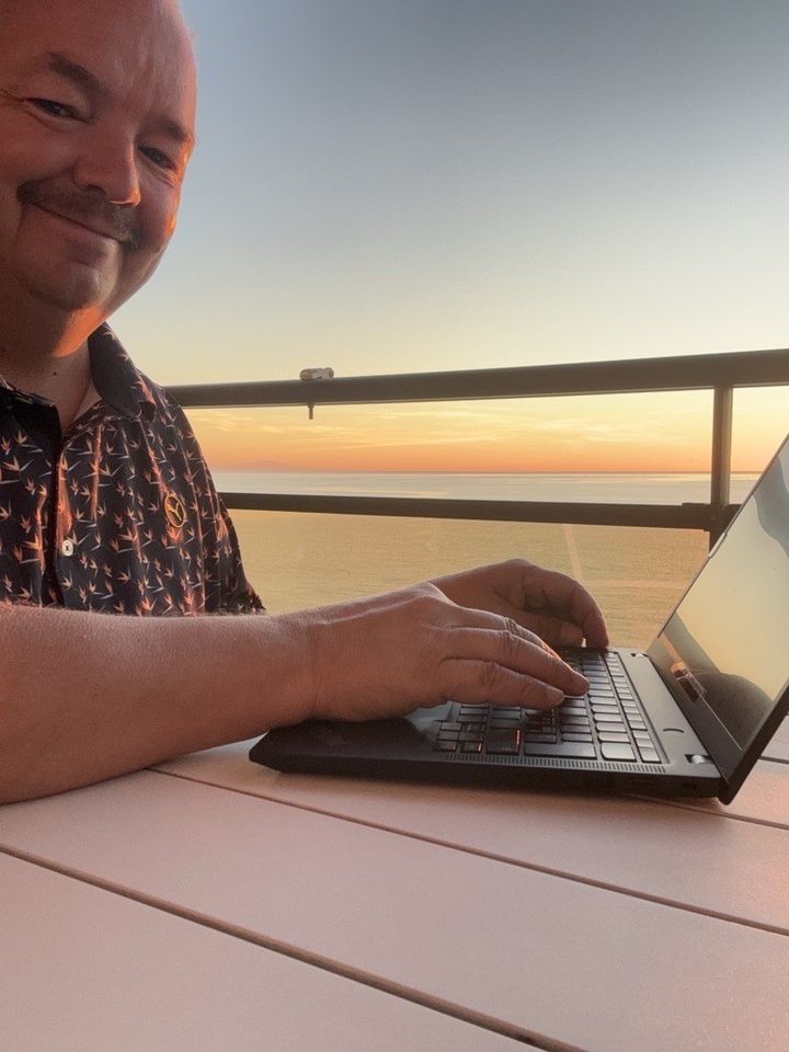 Henkilö katsoo kameraan. Pöydällä läppärin äärellä. Ulkona, taustalla auringonnousu.