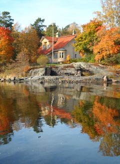 Kuvassa on Villa W. Karsten Helsingissä, jota ympäröivät syksyiset puut. Rakennuksessa on jyrkkä punainen katto, erkkeri, pieniruutuiset ikkunat ja koristeelliset savupiiput. Villa heijastuu vedenpintaan etualalla.
