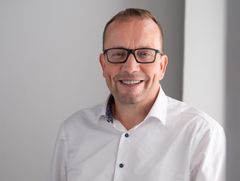 Valokuva henkilöstä, joka hymyilee ja käyttää silmälaseja. Hänellä on valkoinen paita.