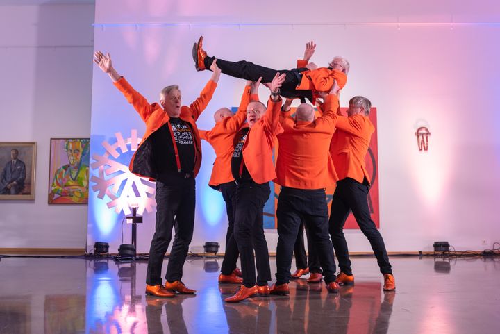 Puumiehet Show tanssijaryhmä tanssii museossa oransseissa puvun takeissa.