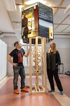 Oliver Kochta-Kalleinen ja Tellervo Kalleinen seisovat puurakenteen ympärillä, johon on kiinnitetty valotaulu.