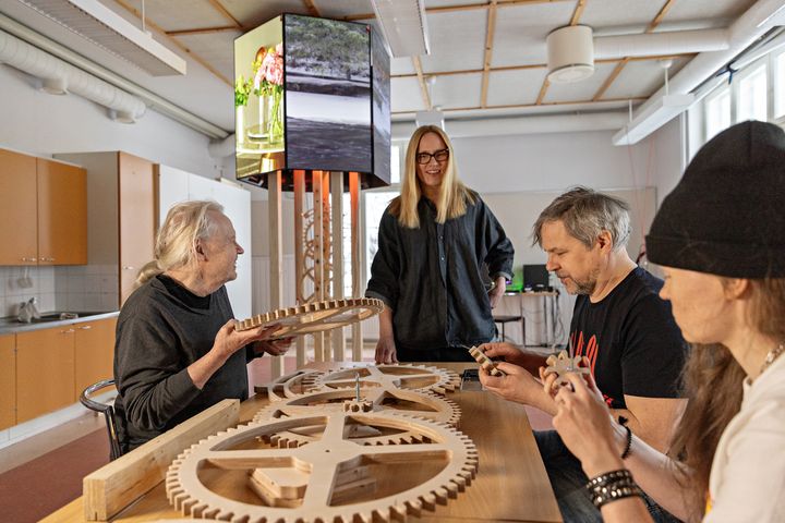 Kuva esittää neljää henkilöä työskentelemässä suurten puisten rattaiden parissa työpajassa.