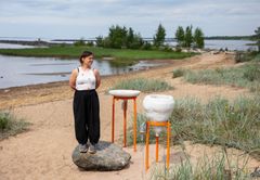 En konstnär står på en sten bredvid sitt verk vid en flodstrand med gräs och buskar.