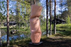 Saviveistoksesta tehty korkea taideteos sijaitsee metsän keskellä lammen rannalla. Taustalla näkyy puita ja rakennuksia.