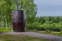 Suuri, pystysuuntainen tervatynnyri seisoo vehreässä maisemassa, taustallaan joki ja koivuja.