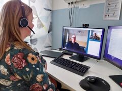 En person sitter med ryggen snett mot kameran och har hörlurar på sig. Framför hen på bordet finns en datorskärm där en annan person syns. De samtalar med varandra.