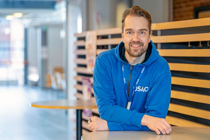 Tuomas Tarkki kuvattuna nojaamassa pöytään. Tuomaksella on päällään OSAOn sininen huppari.