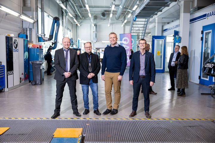 Miehet seisovat autohallissa. Taustalla juhlayleisöä ja OSAOn liput.