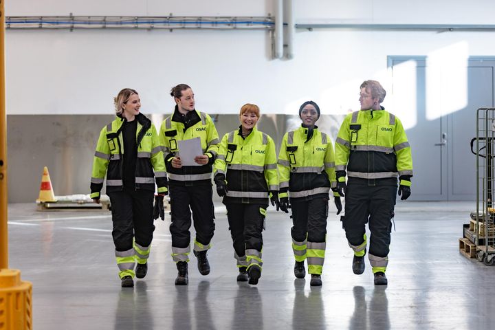 Henkilöt kävelevät rivissä hallissa keltaiset huomioasut päällään.