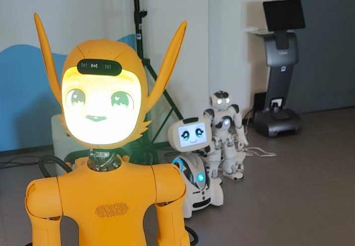 Kuvassa on oranssi humanoidirobotti Miroki ja kaksi muuta robottia RoboHoiva-oppimisympäristössä.