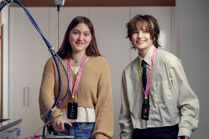 Kaksi tekstiili- ja muotialan opiskelijaa poseeraa hymyillen kameralle silityspöydän ääressä. Molemmilla on kaulassaan värikkäät Kulttuuriagentti-kaulanauhat.