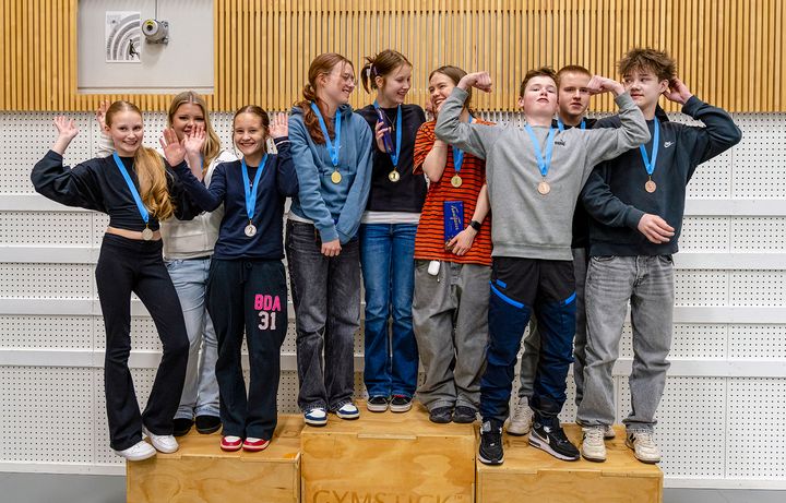 Ryhmä nuoria seisoo korokkeella mitalit kaulassaan. He näyttävät iloisilta ja poseeraavat eri asennoissa.