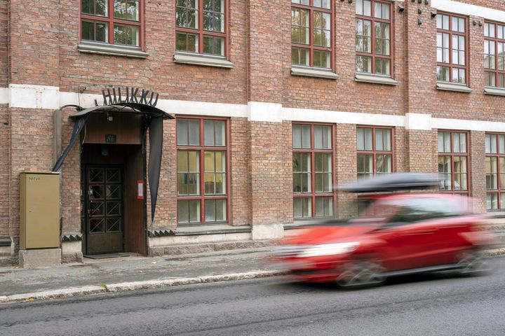 Punainen auto ajaa ohi tiilirakennuksen, jossa on suuria ikkunoita.