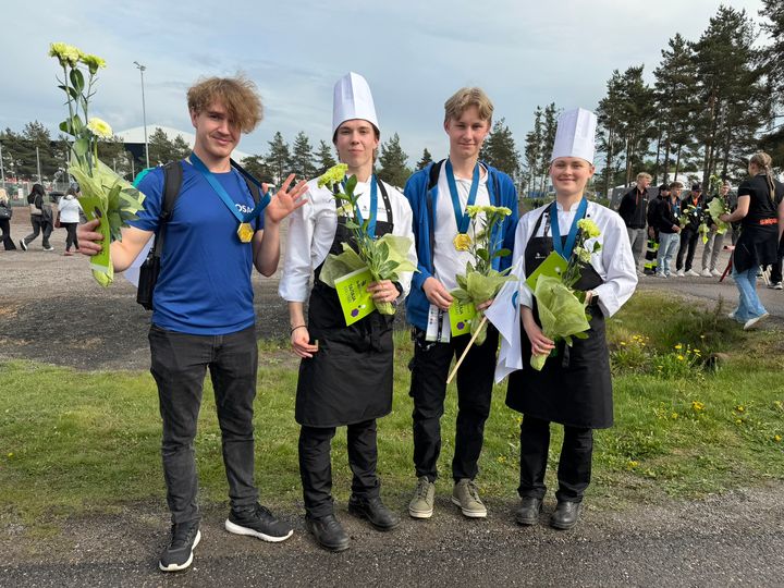 Neljä nuorta seisoo ulkona, kaikilla on kukka kimppu ja mitali kaulassa. Kaksi heistä on pukeutunut kokin takkiin ja hattuun.