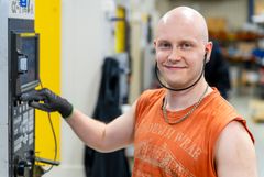 Henkilö tehtaassa, operoimassa koneen ohjauspaneelia, hymyilee kameralle.