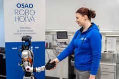 Nainen kättelee robottia laboratoriossa. Taustalla näkyy "OSAO Robohoiva" -mainos.