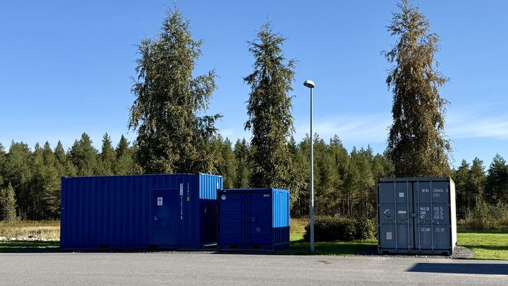 Kolme suurta konttia pysäköitynä ulkoalueelle, taustalla puita ja selkeä taivas.