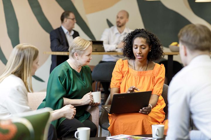 Ryhmä ihmisiä keskustelee yhdessä toimistotilassa. Etualalla naiset keskustelevat kannettavan tietokoneen äärellä.