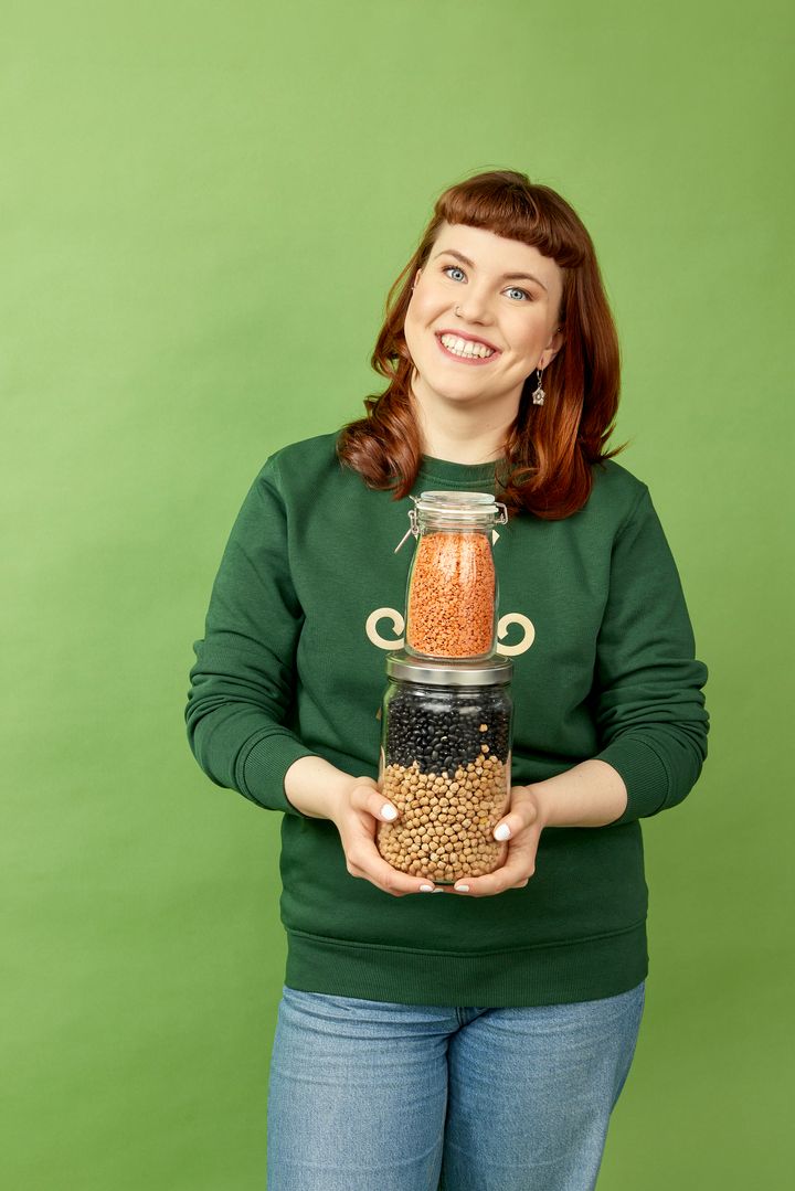 Kuvassa nuori nainen hymyilee ja kädessä kaksi säilykepurkkia, joissa papuja ja linssejä.