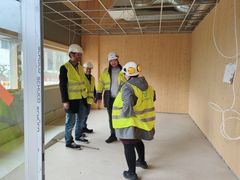 Kuvassa neljä henkilöä rakennustyömaalla keltaisissa huomioliiveissä ja suojakypärissä keskustelemassa puisen seinän edessä.