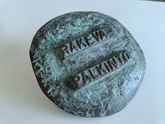 Pöydällä pidettävä, pyöreä, vihertävänpatinoitunut veistos, jossa kohokirjaimin teksti "RAKEVA-PALKINTO".