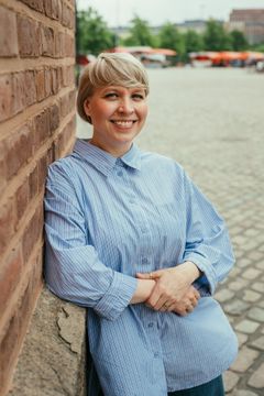 Nainen seisoo ulkona tiiliseinään nojaten.