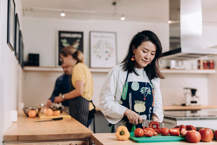 Nainen leikkaa vihanneksia keittiössä, taustalla kaksi henkilöä työskentelee.