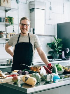 Mies seisoo nykyaikaisessa keittiössä leveästi hymyillen, edessään valikoima raakoja vihanneksia ja hedelmiä.