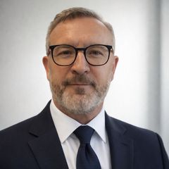 Kuvassa on liikemies, jolla on harmaa tukka ja parta, yllään puku ja silmälasit, seisomassa neutraalilla taustalla.