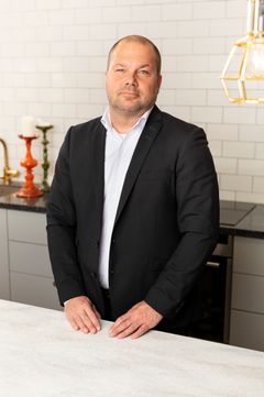 Mies seisoo keittiössä pukeutuneena mustaan pukuun ja valkoiseen paitaan.