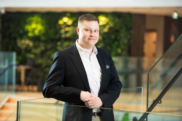 Tuomas Salminen kuvattuna Ilmalan aulassa: taustalla vaaleaa puuta ja vihreää. Salminen hymyilee kuvassa.