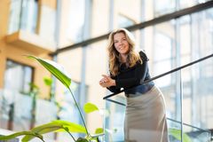 Woman smiling and leaning on a glass railing inside a modern building with plants in the foreground.