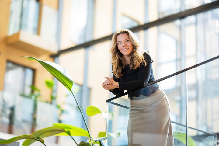 Woman smiling and leaning on a glass railing inside a modern building with plants in the foreground.