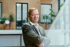 Hymyilevä keski-ikäinen mies puvun takissa, kädet puuskassa rinnan päällä, taustalla okrankeltainen ikkunaseinä