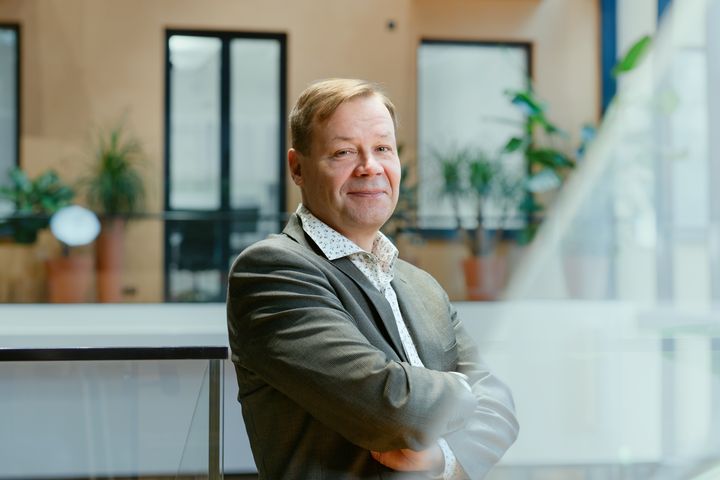 Hymyilevä keski-ikäinen mies puvun takissa, kädet puuskassa rinnan päällä, taustalla okrankeltainen ikkunaseinä