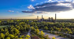 Ilmakuva, joka esittää vehreää puistomaista maisemaa etualalla, taustalla teollisuutta. Etualan puistossa on vihreitä puita ja nurmialueita, ja taustalla näkyy savupiippuja ja teollisuusrakennuksia, horisontissa kaupunki auringon paisteessa.