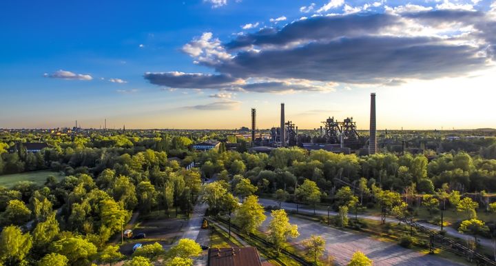 Ilmakuva, joka esittää vehreää puistomaista maisemaa etualalla, taustalla teollisuutta. Etualan puistossa on vihreitä puita ja nurmialueita, ja taustalla näkyy savupiippuja ja teollisuusrakennuksia, horisontissa kaupunki auringon paisteessa.