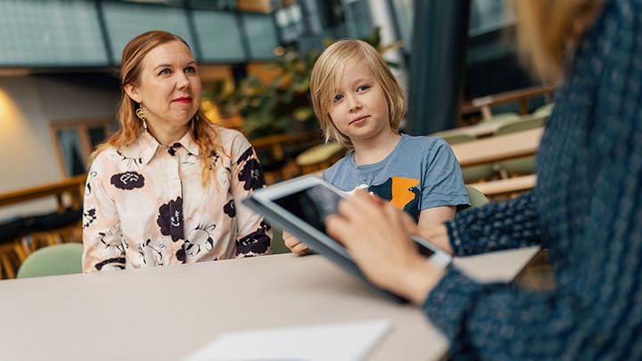 Sosiaalihuollon ammattilainen tabletilla, taustalla asiakkaat, äiti ja lapsi