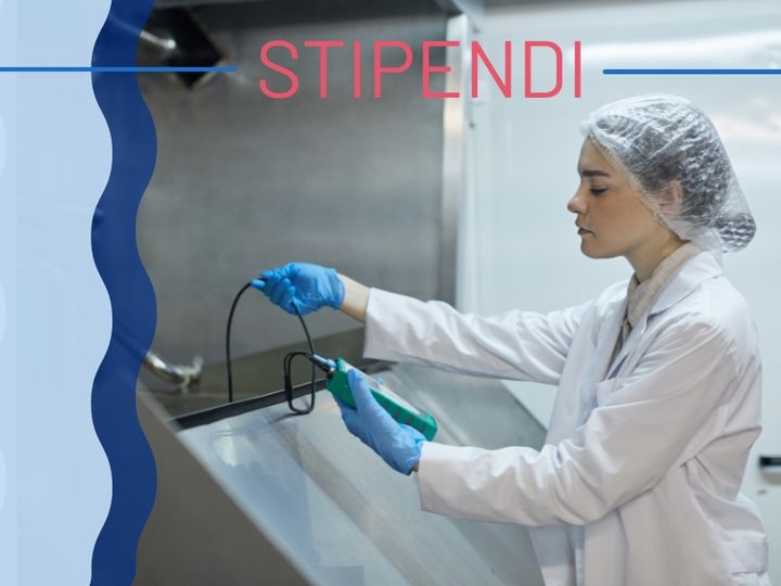 Nainen laboratoriotakissa ja suojahatussa työskentelee tutkimuslaitteiston parissa. Teksti "STIPENDI" ylhäällä.